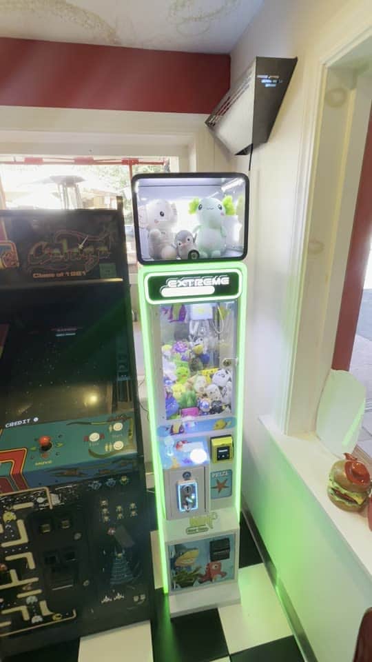 A green-lit claw machine beside arcade cabinets on a checkered diner floor