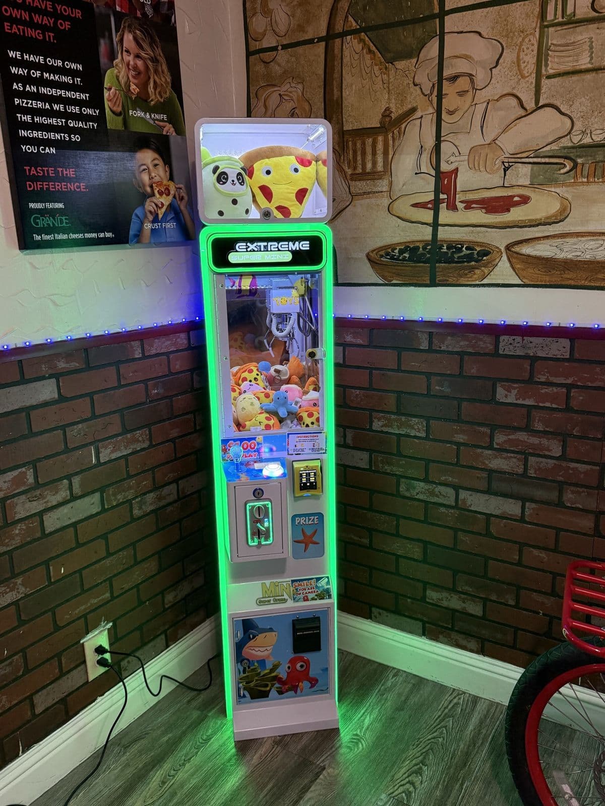 A green-lit California Claw machine in the corner of a pizzeria, brick wall and mural behind it