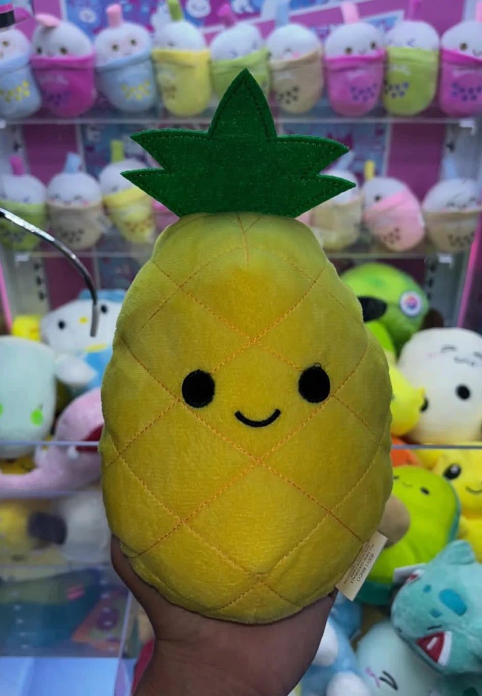A hand holding a smiling pineapple plushie won from a California Claw machine, with rows of boba-cup plushies behind it