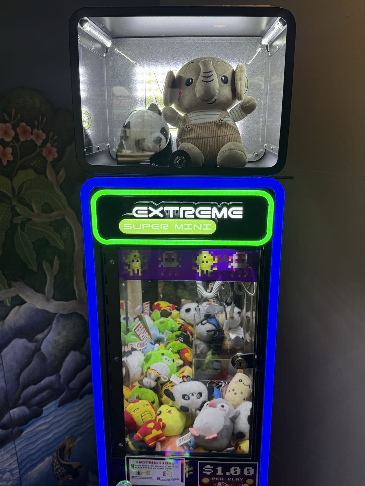 A blue-lit California Claw machine against a waterfall mural, an elephant plush on top